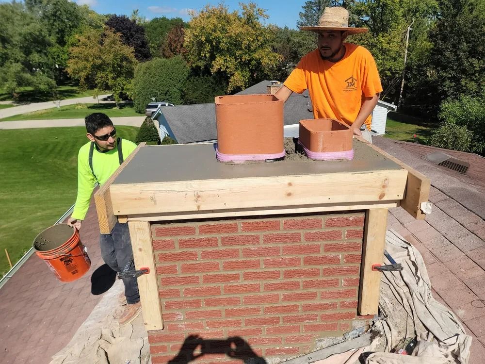 Two workers repair a brick chimney on a rooftop, with mortar and chimney pots.
