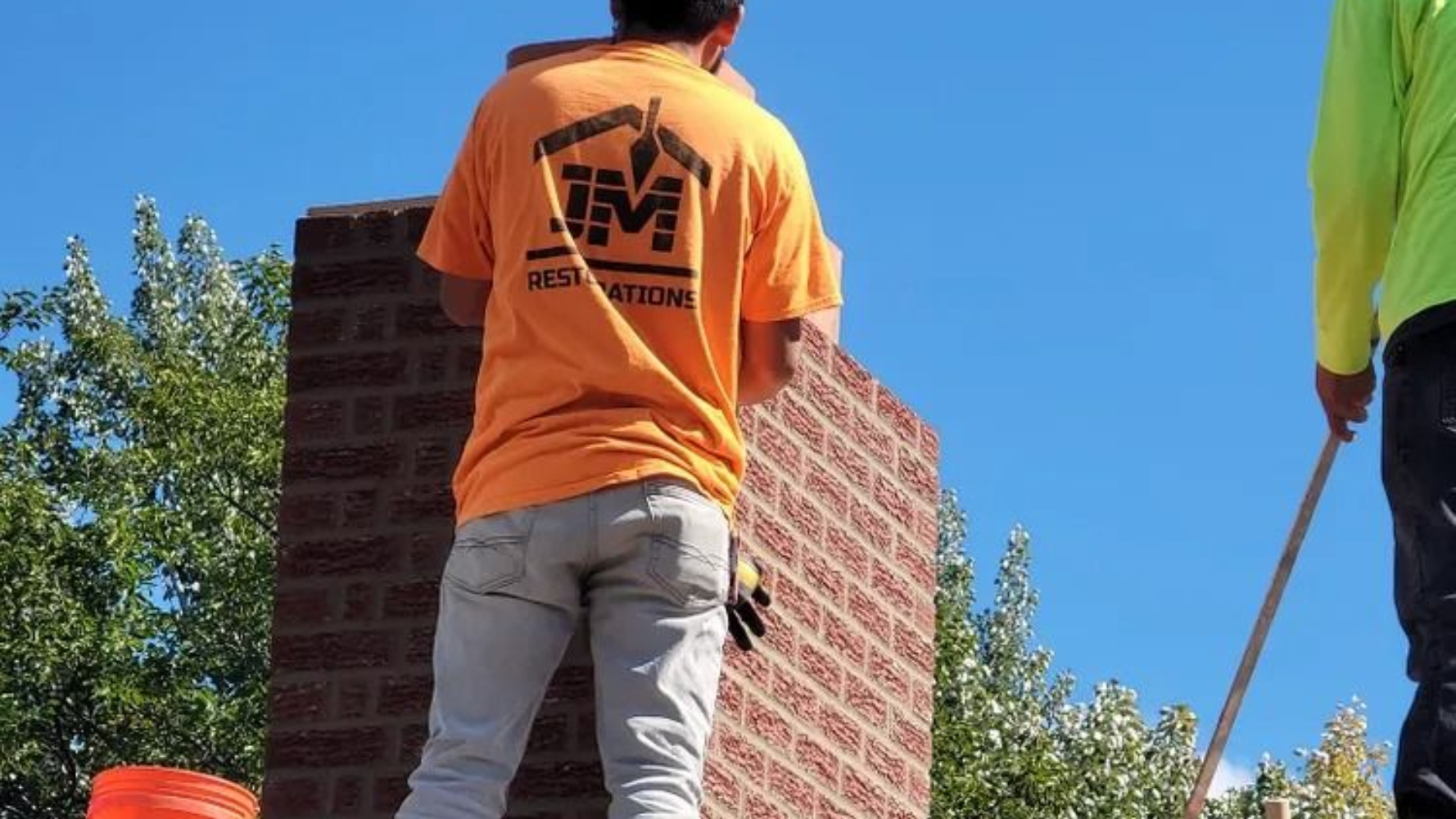 Two workers in orange and blue shirts building a brick wall outdoors under a clear sky