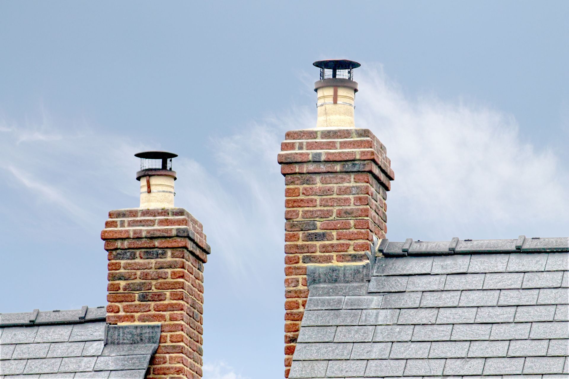 Two brick chimneys with metal caps rising above gray rooftops against a blue sky.