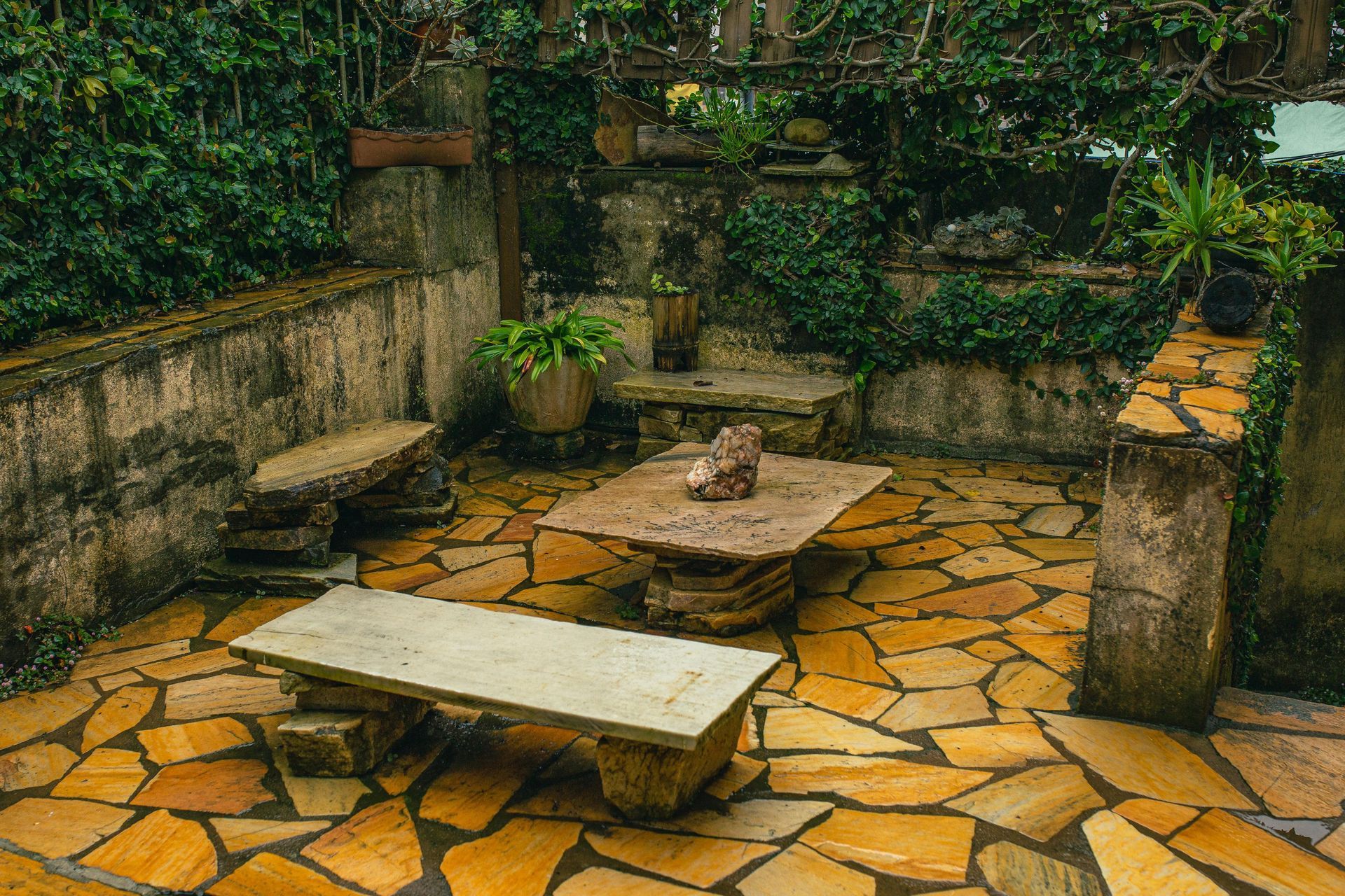 Stone patio with low benches and a square table, surrounded by potted plants and ivy-covered walls