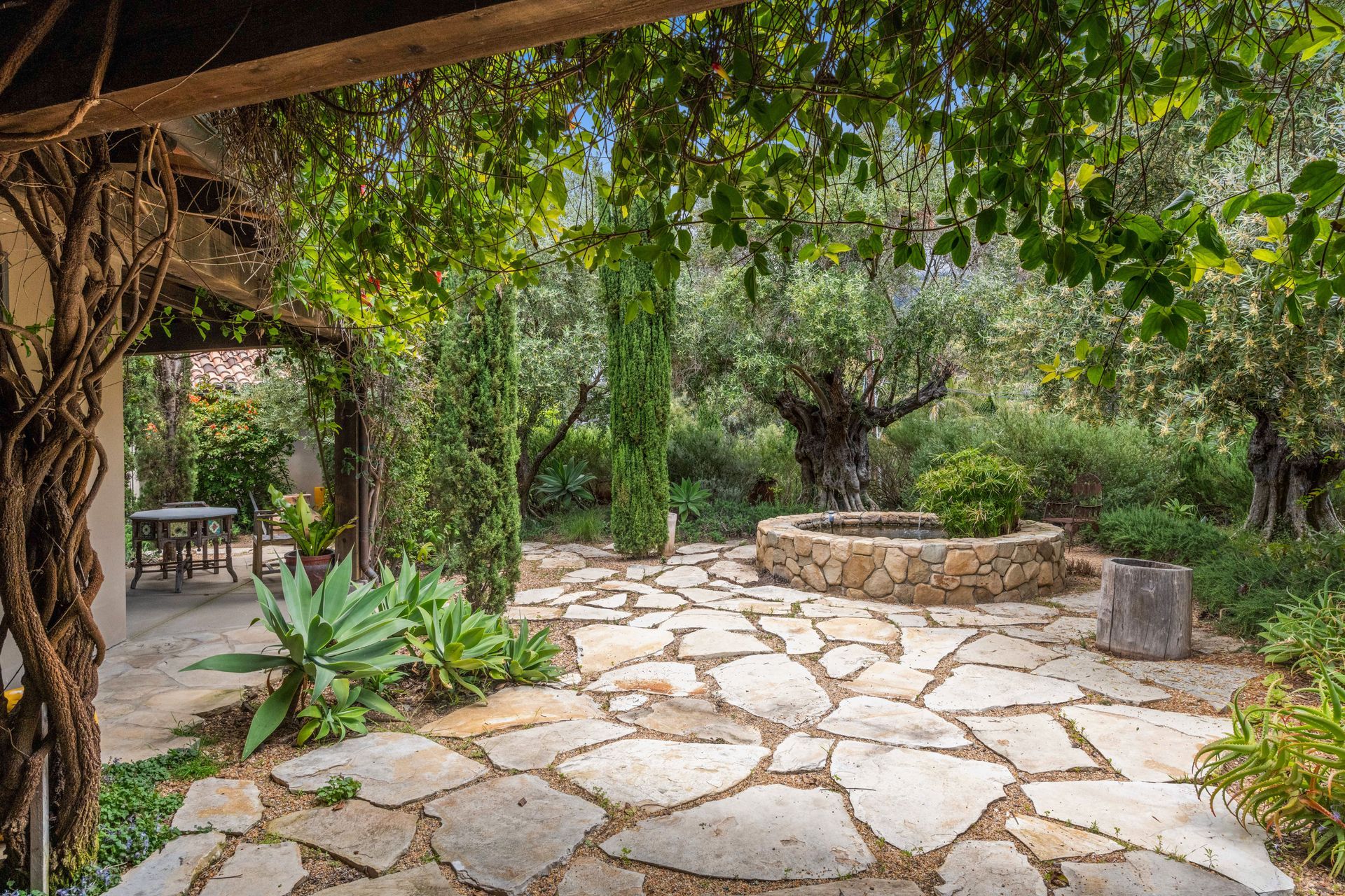 Shaded stone patio with lush greenery, a central round planter, and rustic wooden posts