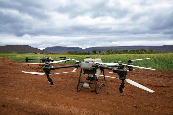 Two drones parked on a dirt field under cloudy skies, with green farmland and hills in the background.