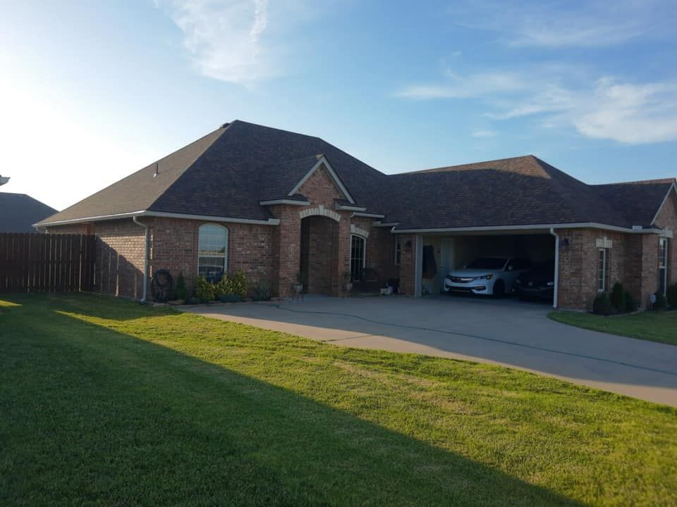 Brick house with attached carport, driveway, and green lawn under a blue sky. Cars are parked in the carport.