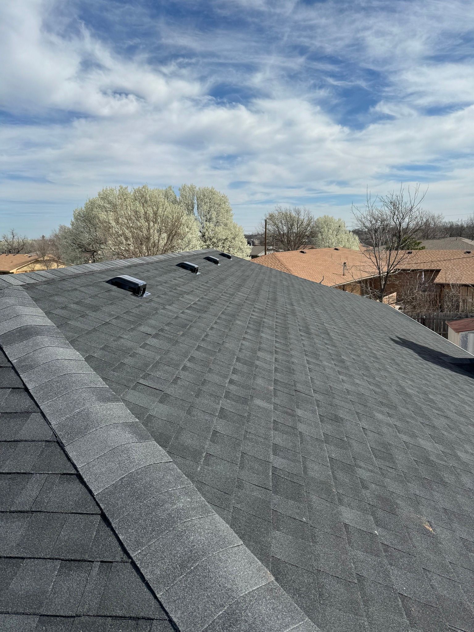 Gray asphalt shingle roof on a sunny day with blue sky and some clouds, view from above.
