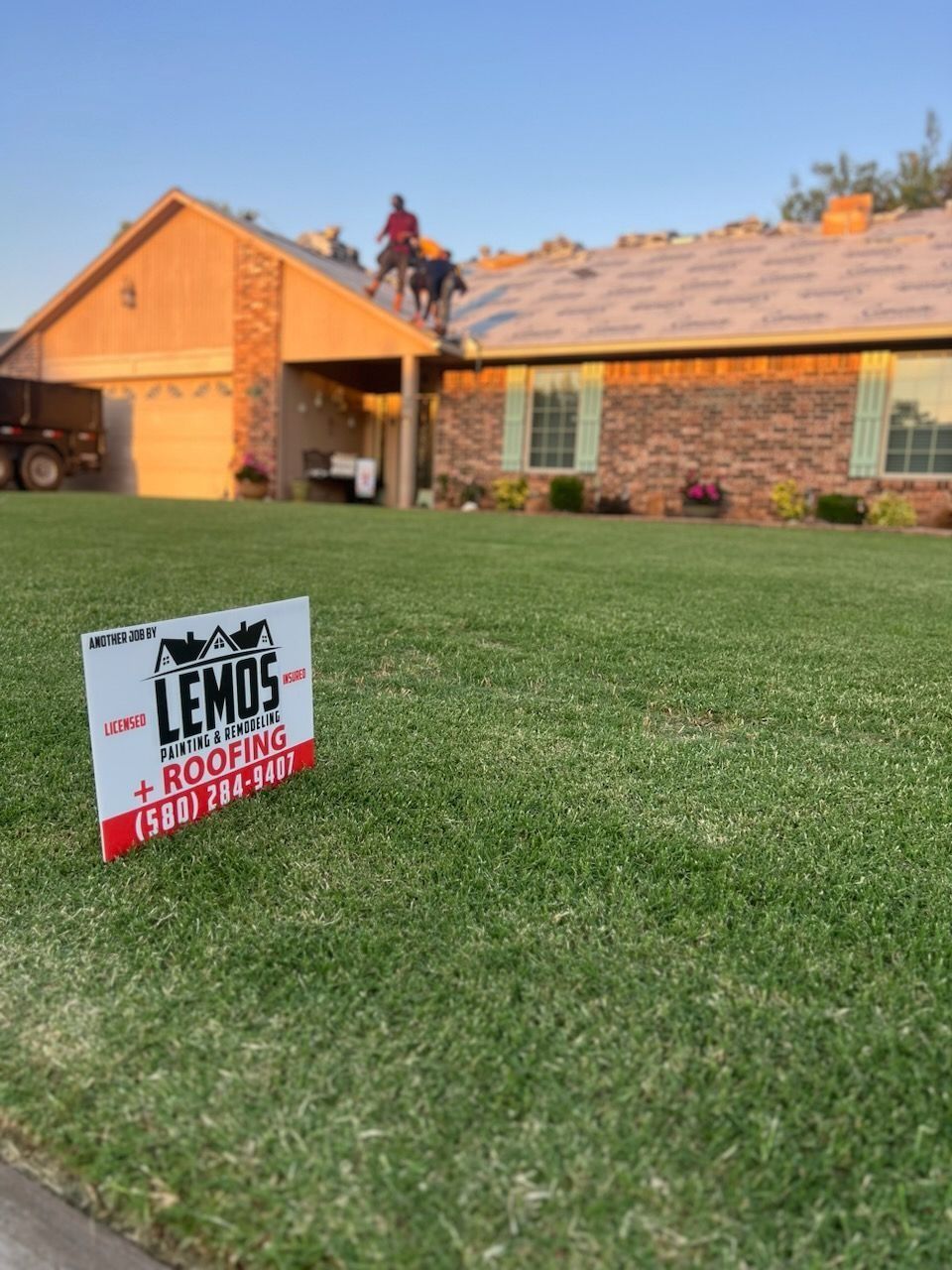 Sign for Lemos Roofing on a lawn; workers on a roof, house in background.