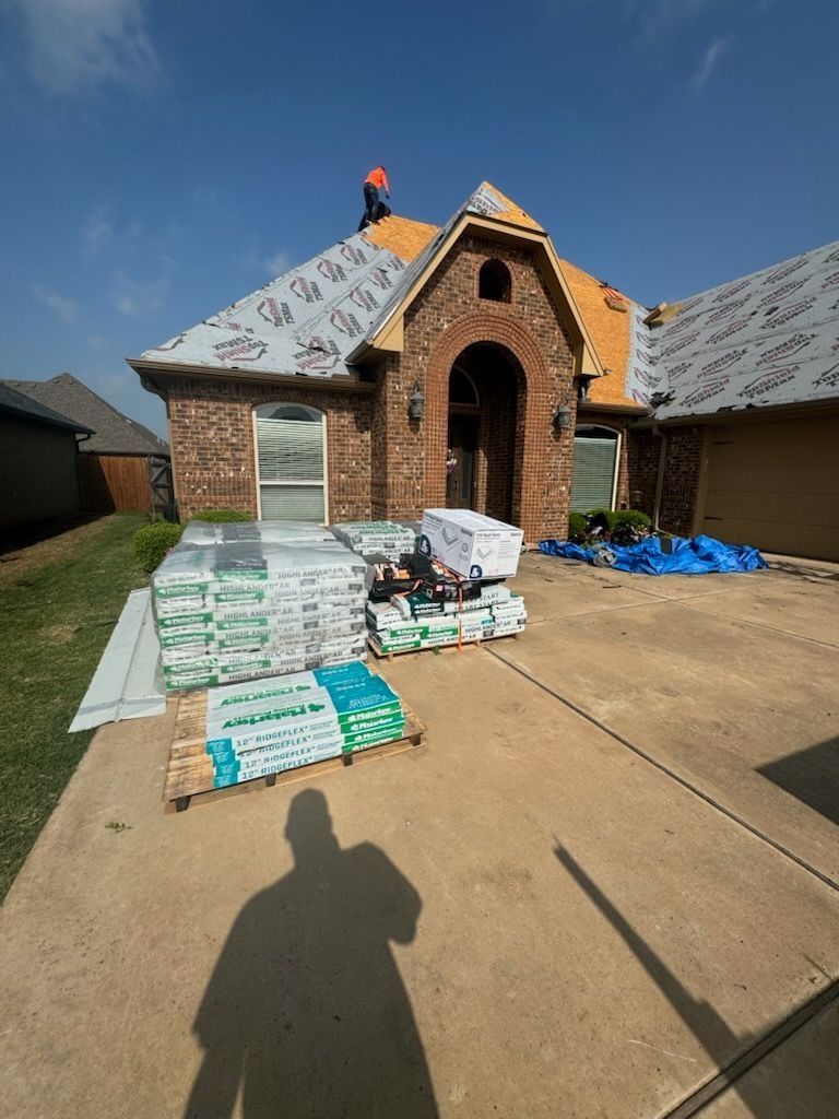 House under construction; roofing materials on driveway. Roofer on the roof, blue tarp, sunny day.