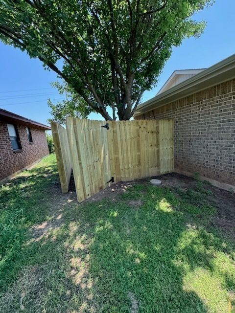 A new wooden fence built beside a brick building and a grassy yard with a large tree.