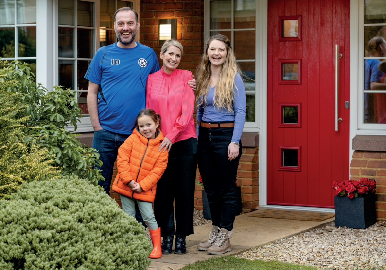 Family of four standing in front of a red front door, posing for a photo.