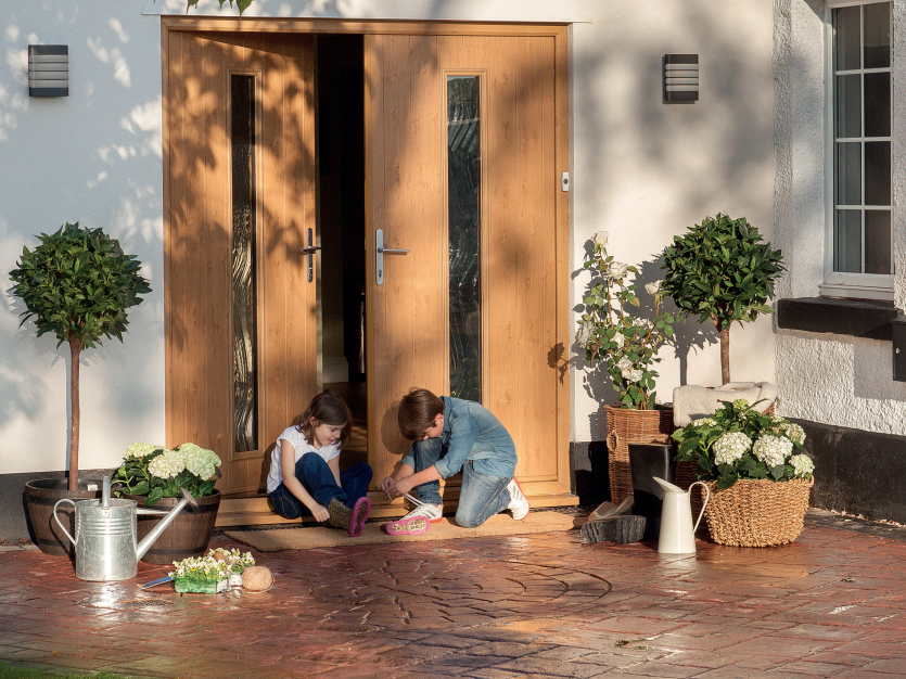 Two children play on a porch in front of a double door, surrounded by potted plants and a watering can.