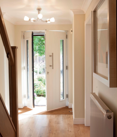Brightly lit entry hall with open white door, revealing green foliage outside. Wooden floors, a radiator, and stairs.