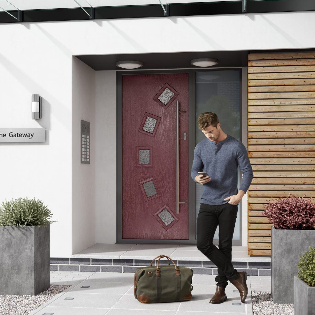 Man leaning against burgundy door, using phone. A green bag sits at his feet. Modern house exterior.