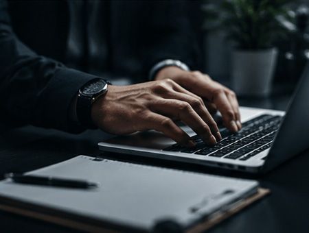 Hands typing on a laptop beside a notepad in a dim office setting