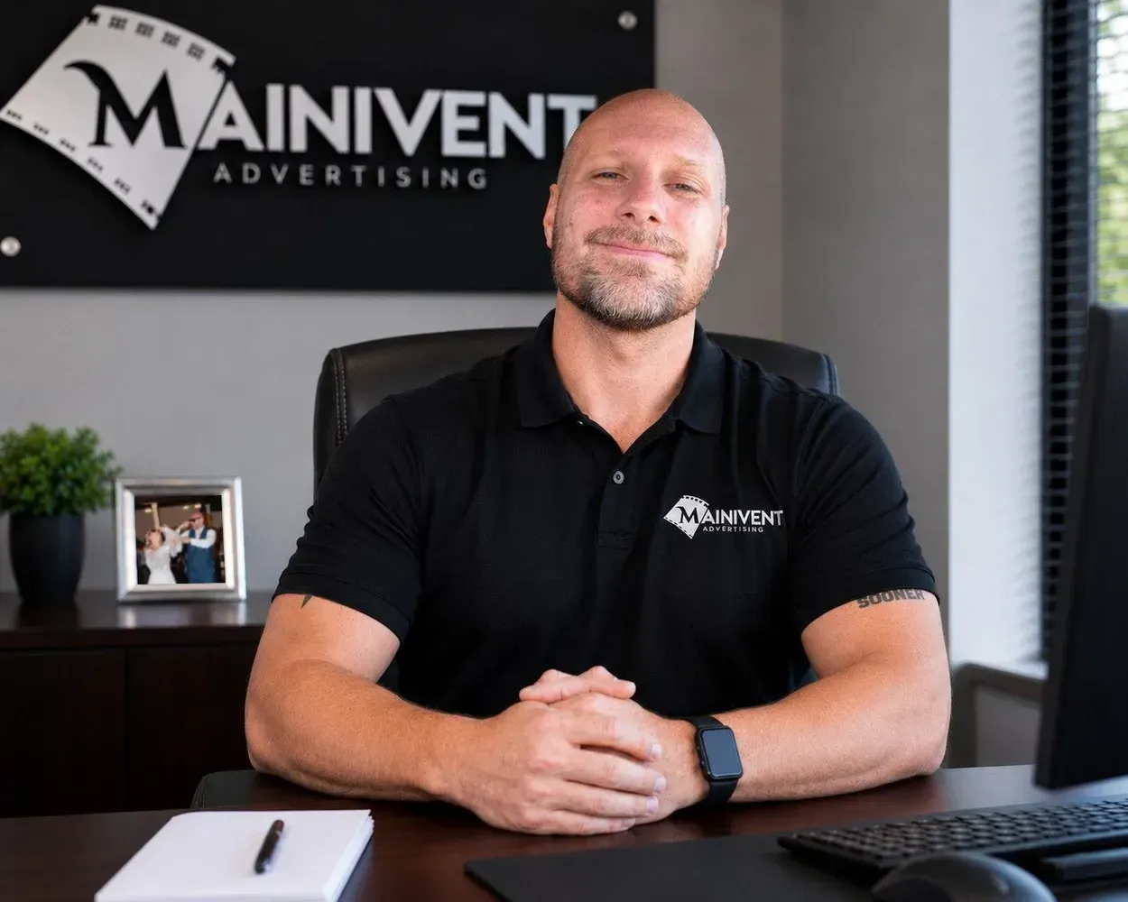 Man sitting at a desk in an office with a Mainvent Advertising sign behind him