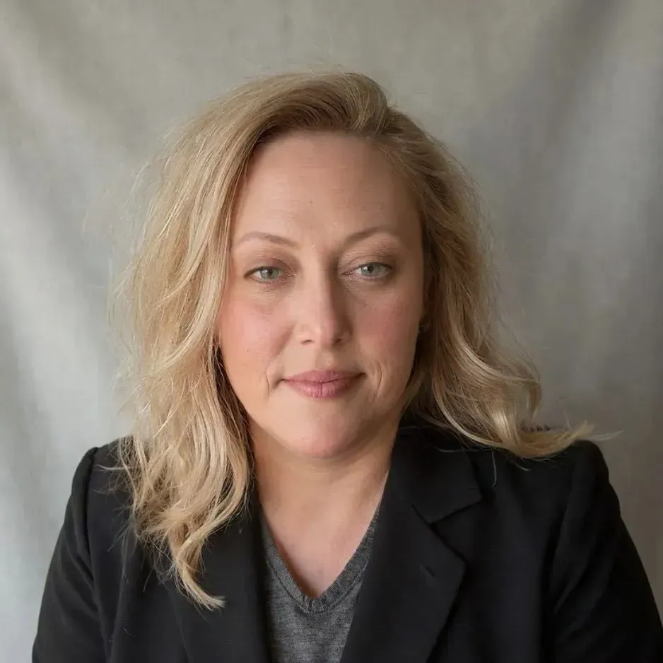 Portrait of a person with blonde hair in a black blazer against a gray background