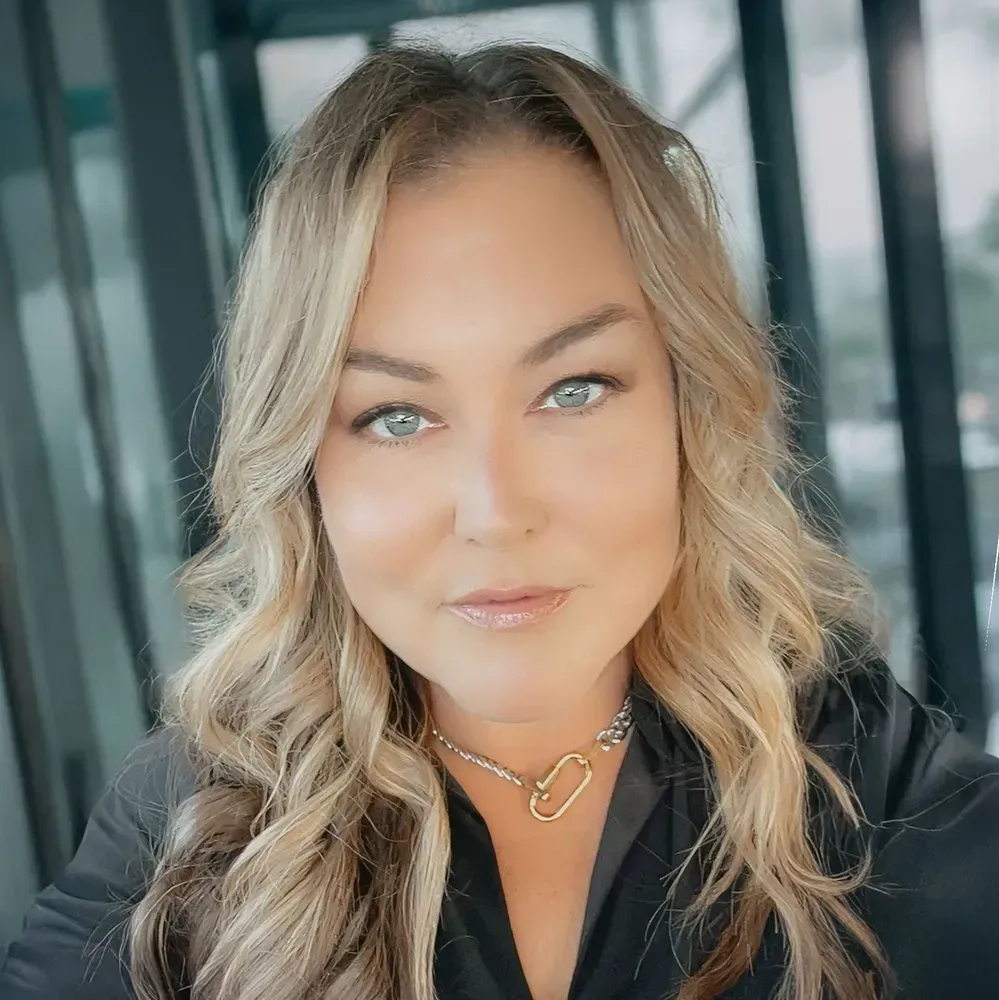 Close-up portrait of a person with wavy blonde hair and blue eyes, wearing a black top and gold necklace.