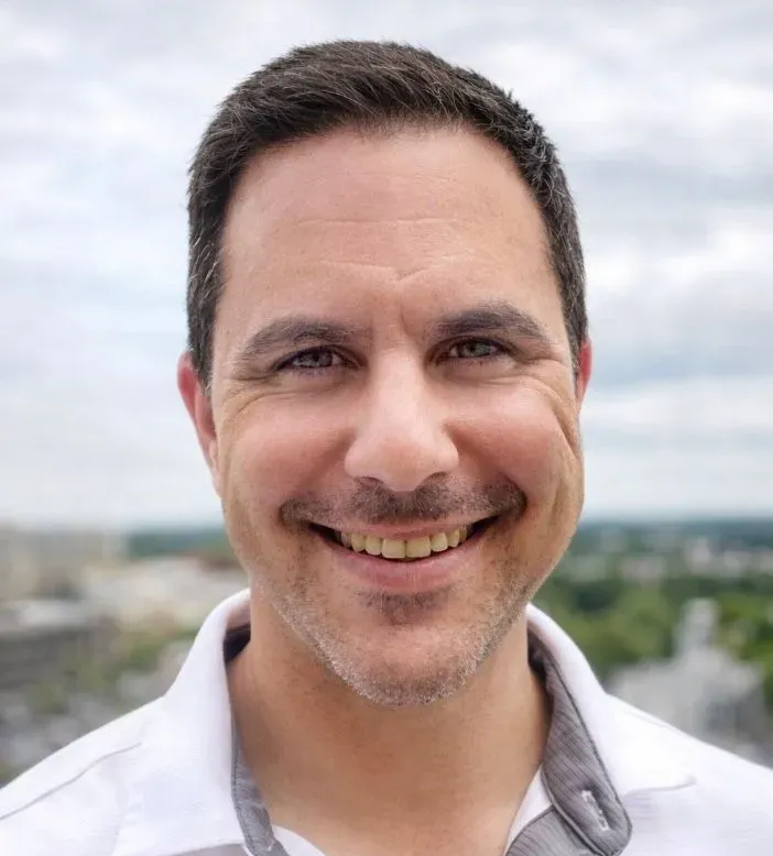 Smiling man in a white shirt outdoors with blurred cityscape and cloudy sky behind him