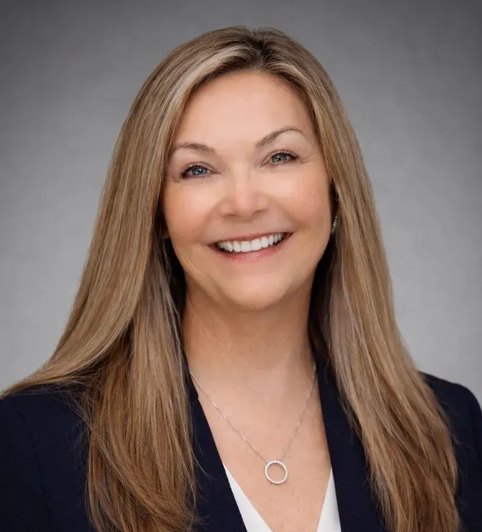 Smiling woman with long blonde hair in a dark blazer, against a gray studio background