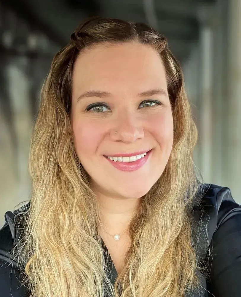 Smiling woman with long wavy blonde hair in a dark top, seated in a softly lit indoor setting.