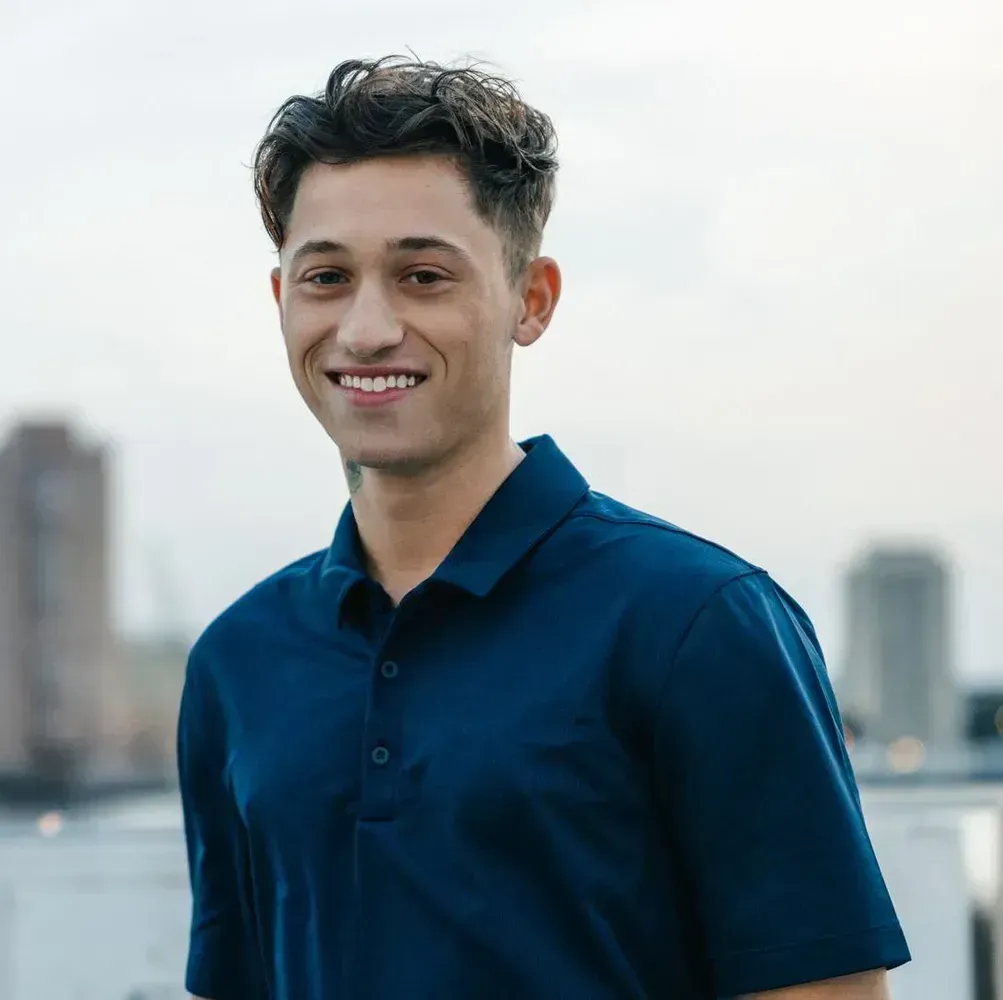 Smiling person in a navy shirt standing outdoors with a blurred city skyline behind them