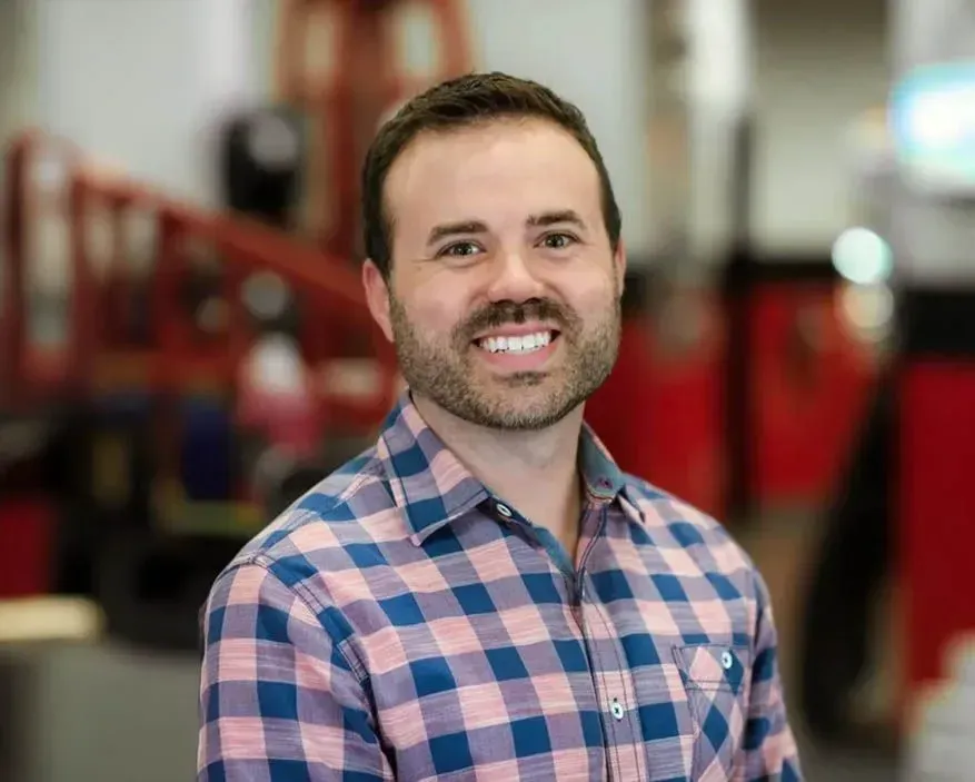 Smiling man in a blue and pink plaid shirt standing in a warehouse with red equipment in the background