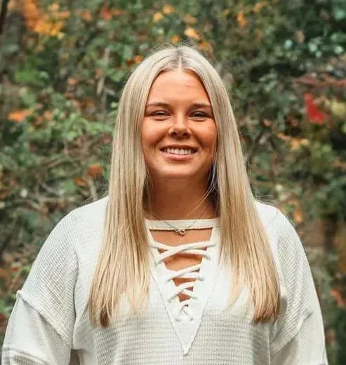 Smiling woman in a white lace-up sweater standing outdoors with blurred greenery behind her