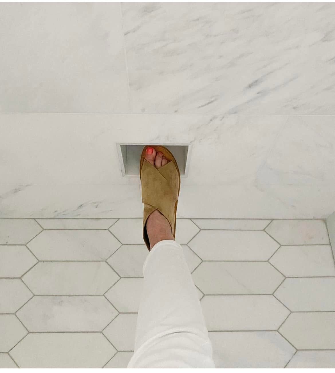 Foot in a tan sandal stepping into a square wall opening above white hex tile floor.