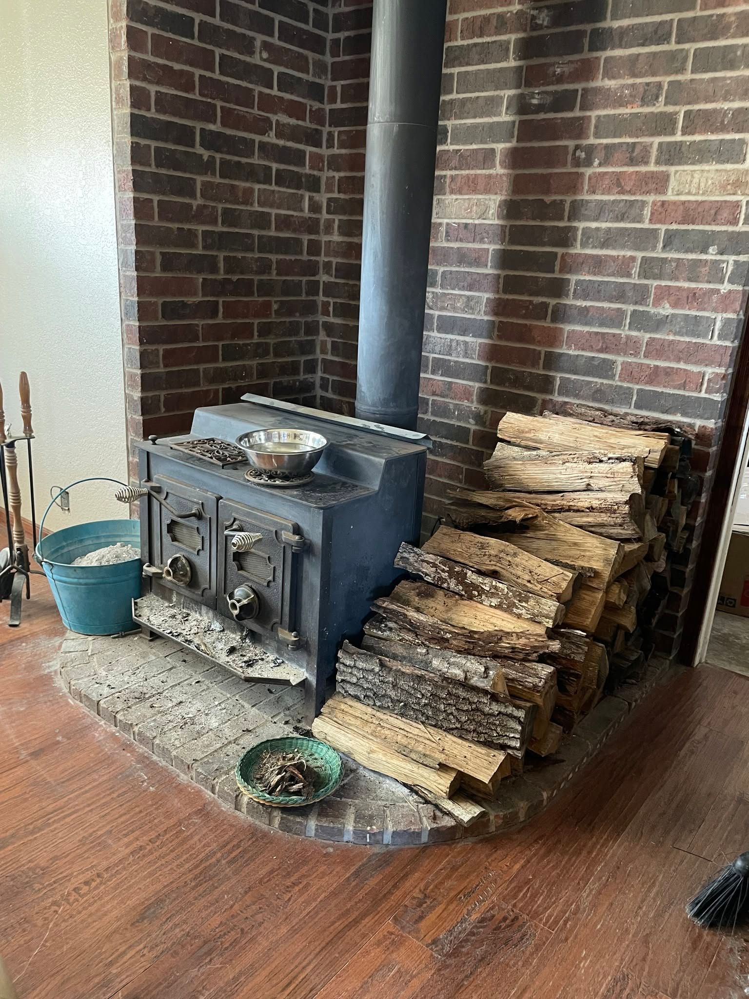 Wood stove in a brick hearth beside stacked firewood and a blue bucket on a hardwood floor