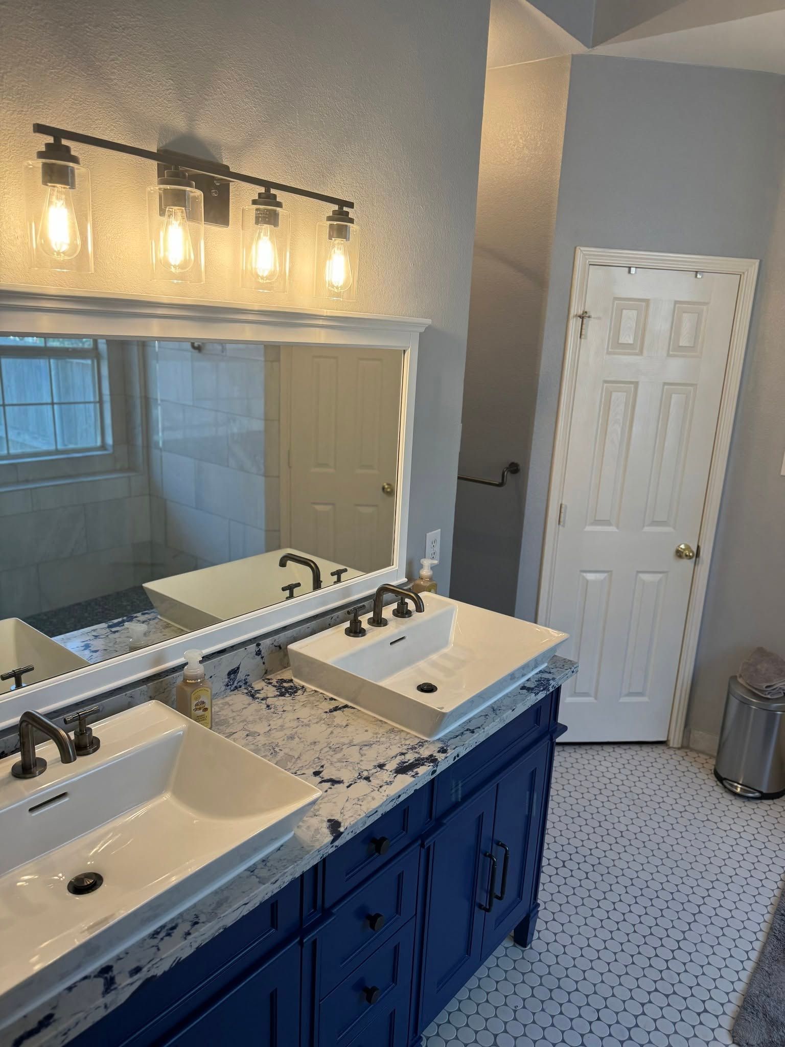 Bathroom with blue vanity, white sink, large mirror, and lit vanity lights beside a shower and closet door.