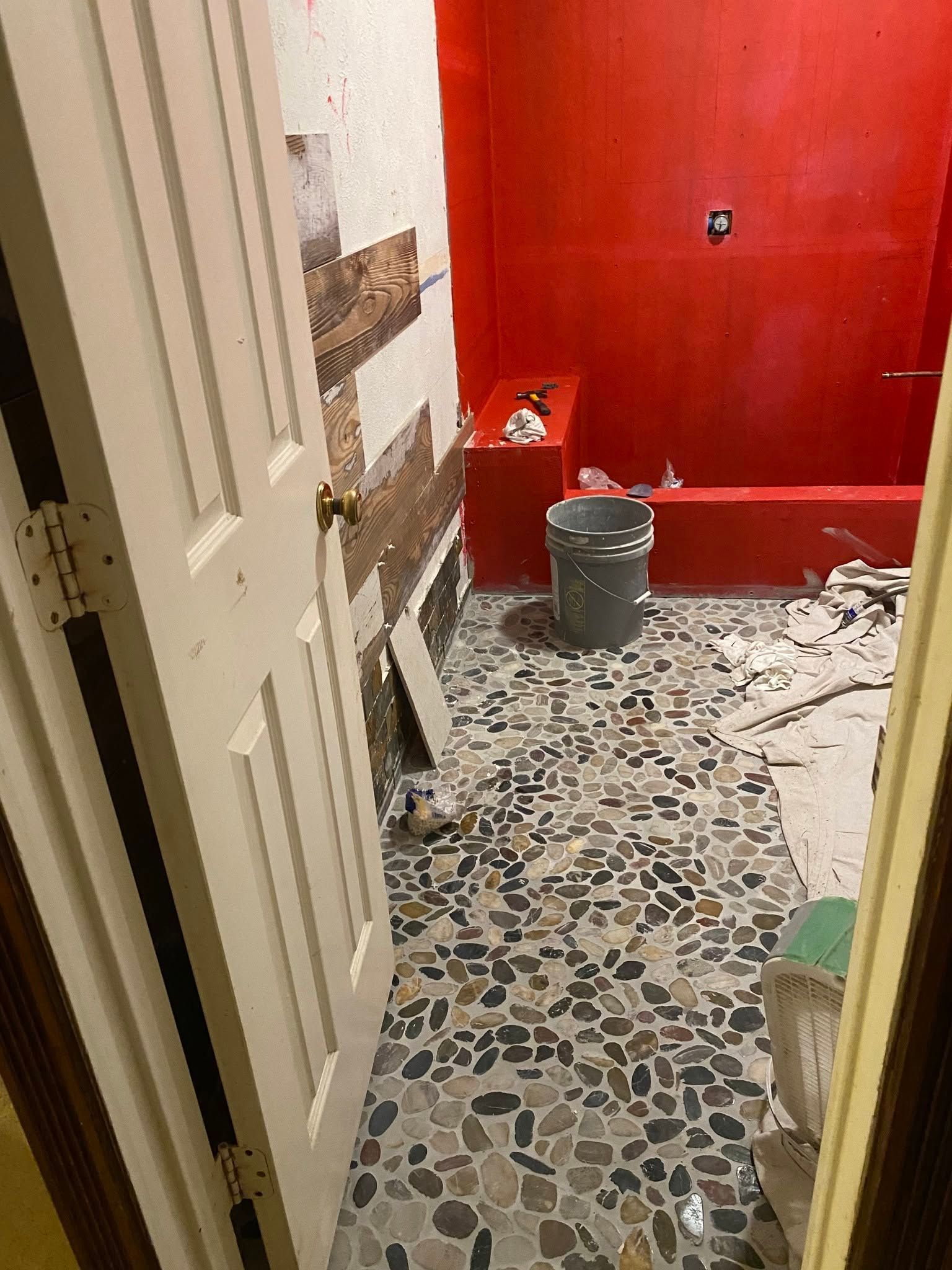 Messy bathroom with red walls, debris on the floor, open door, and a trash can near the sink area