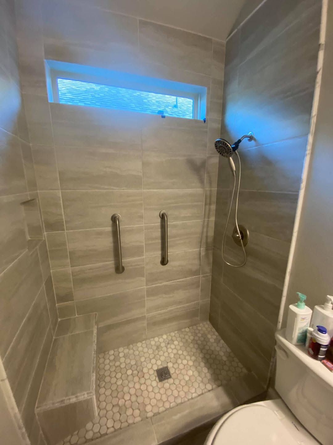 Modern walk-in shower with tiled walls, pebble floor, grab bars, and handheld showerhead.