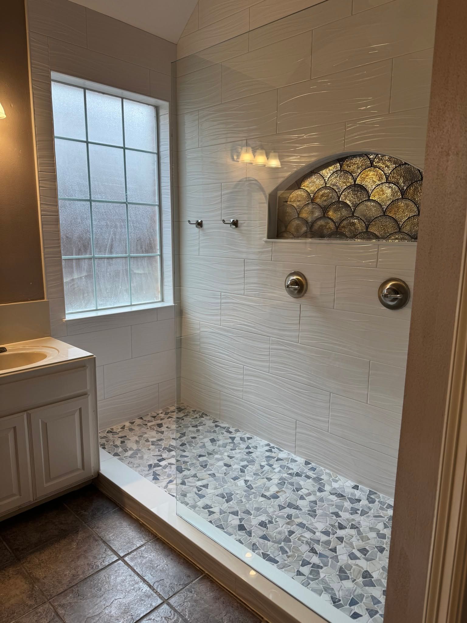 White tiled walk-in shower with mosaic floor, bench, niche, and frosted window beside a vanity.