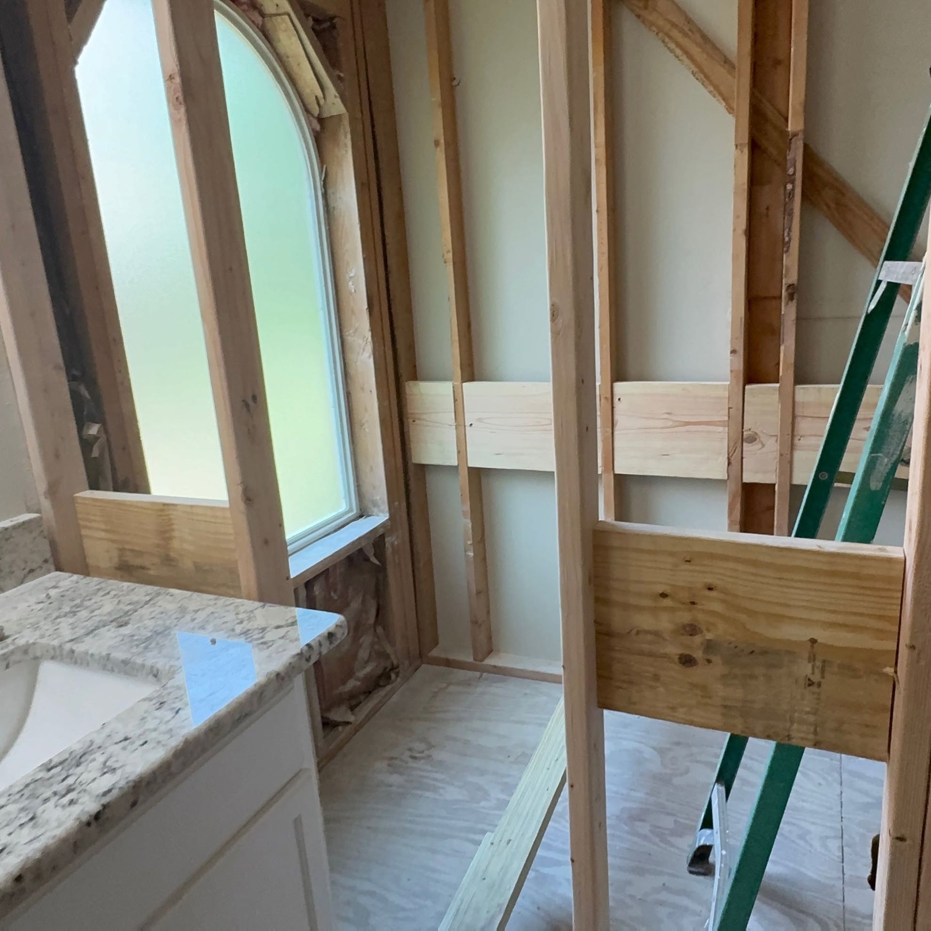 Partially renovated bathroom with exposed wall framing, arched window, and ladder beside a shower area