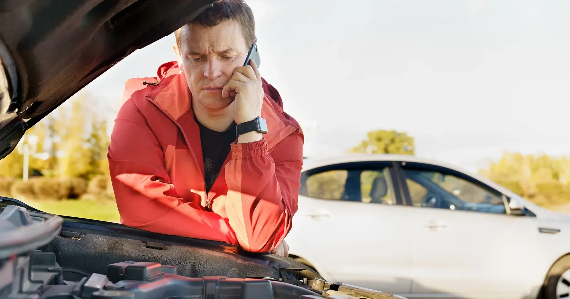 A man is talking on a cell phone while looking under the hood of a broken down car.