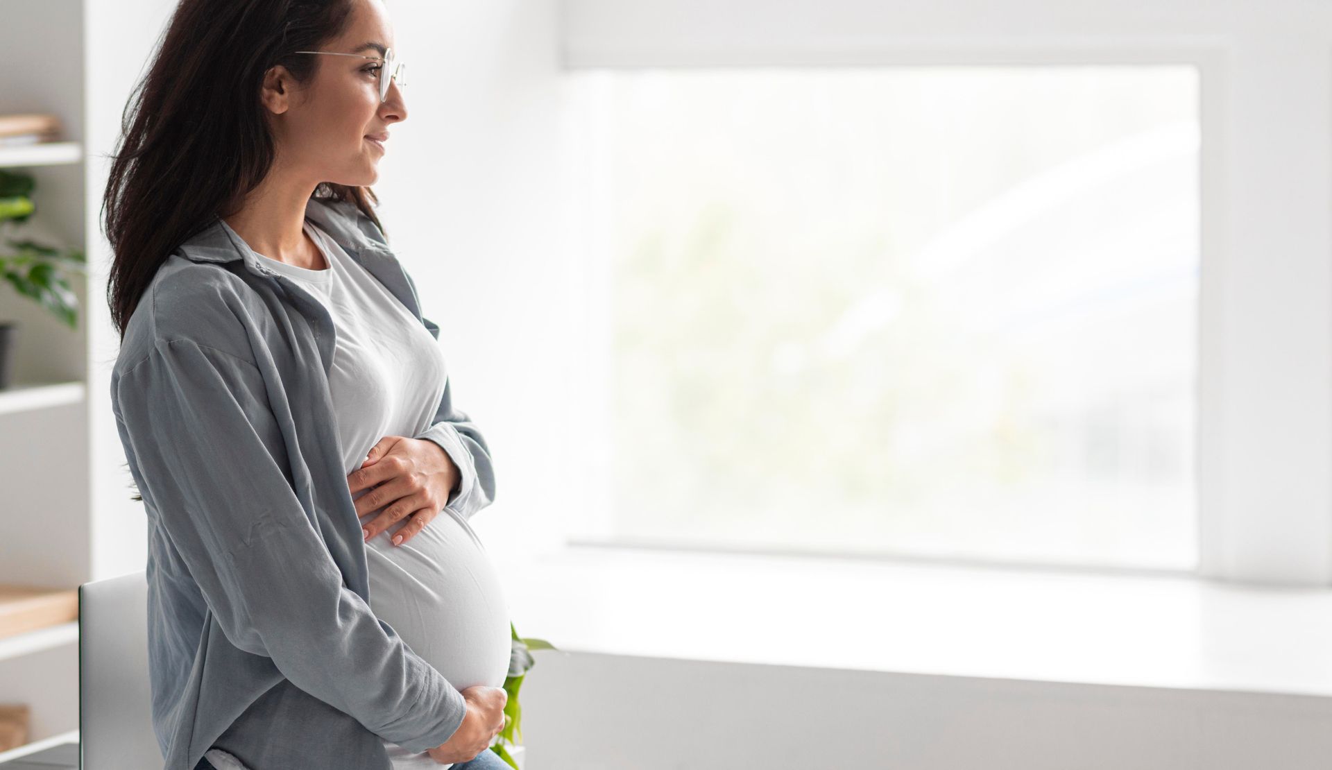 Embracing her pregnancy transformation, a woman stands by a window, holding her belly and smiling. 