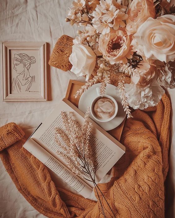 Um livro, uma xícara de café, flores e um suéter estão em uma cama.