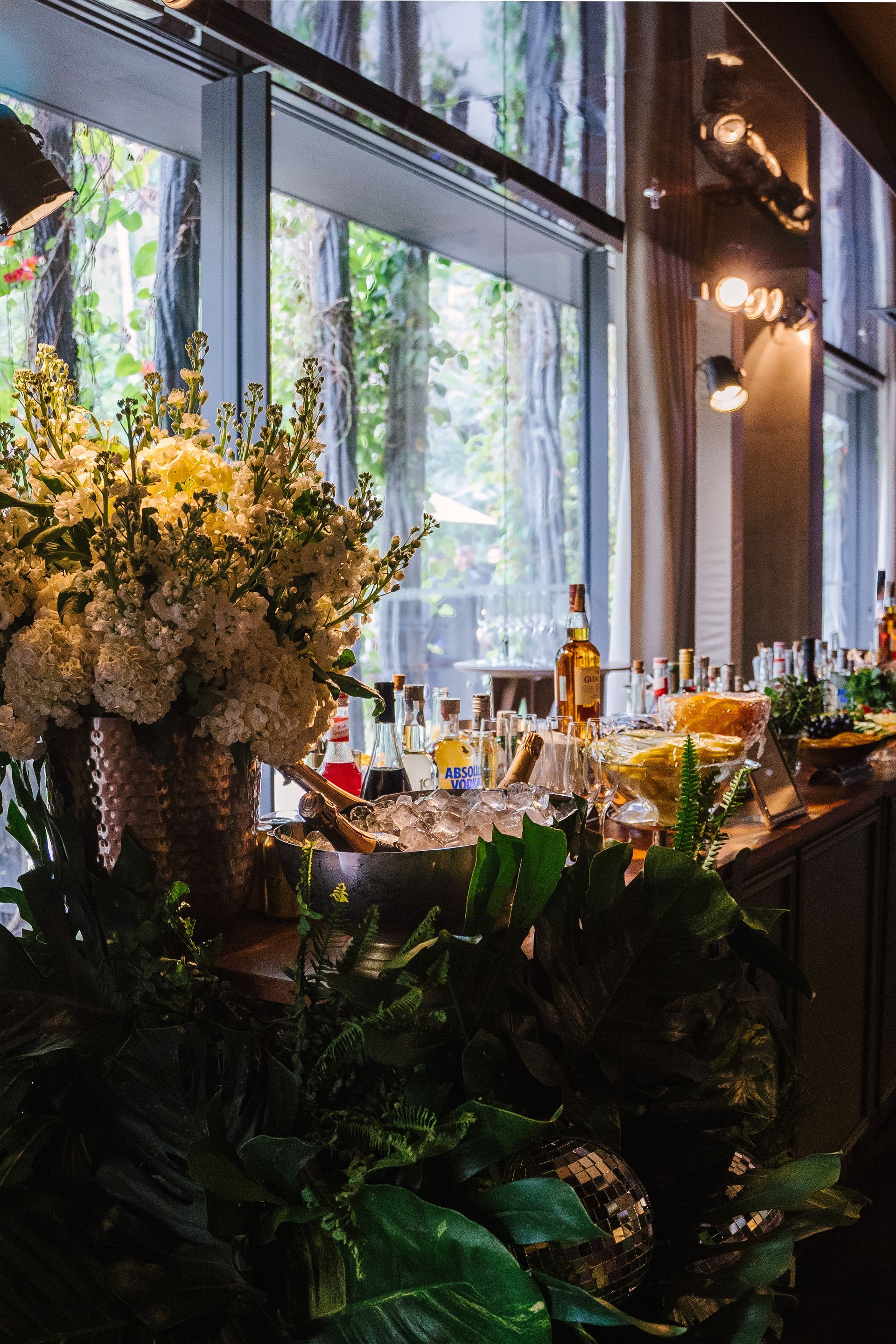 Um bar cheio de garrafas e flores em frente a uma janela.