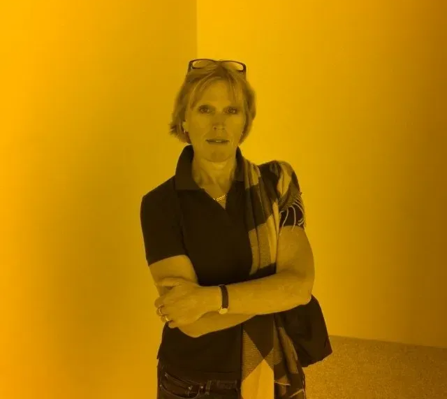 Person standing with arms crossed in a yellow-lit room, wearing a black top and carrying a bag.Elvira Wilthagen
