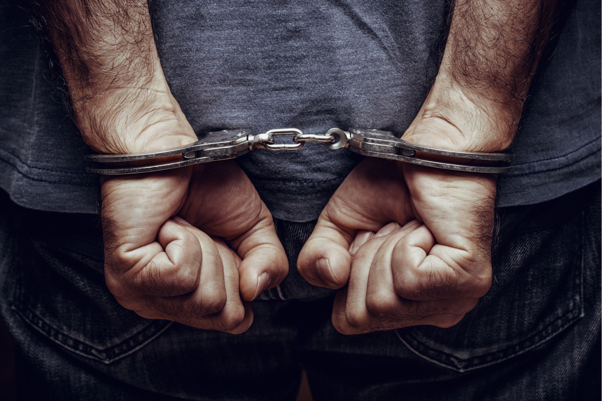 Person's hands cuffed behind their back; wearing jeans and a gray shirt.
