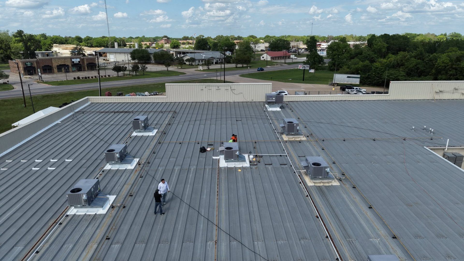 Workers on a gray rooftop inspecting HVAC units under a partly cloudy sky.