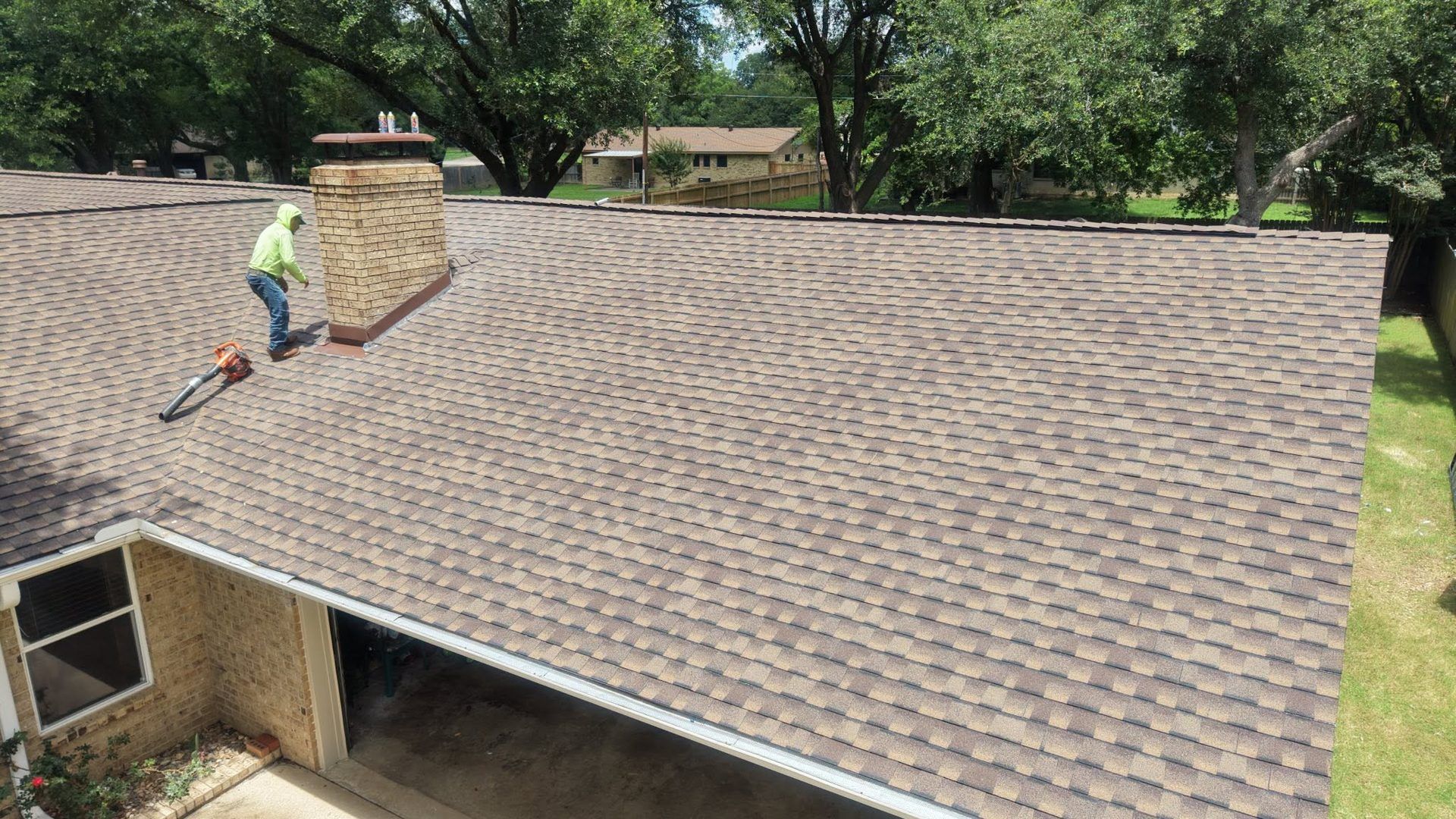 Worker cleaning a large shingled roof with a leaf blower near a house and trees