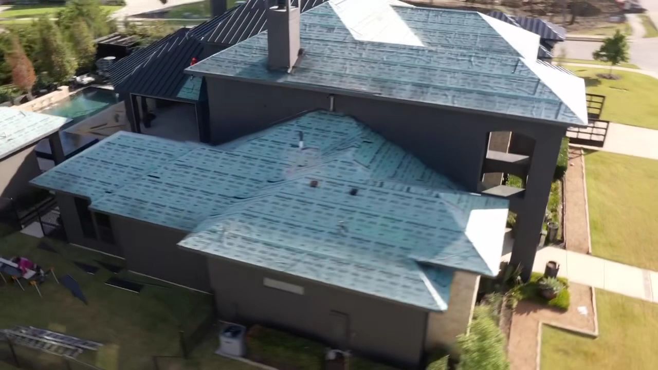Aerial view of a house with a teal roof and chimney, surrounded by a lawn and sidewalk.