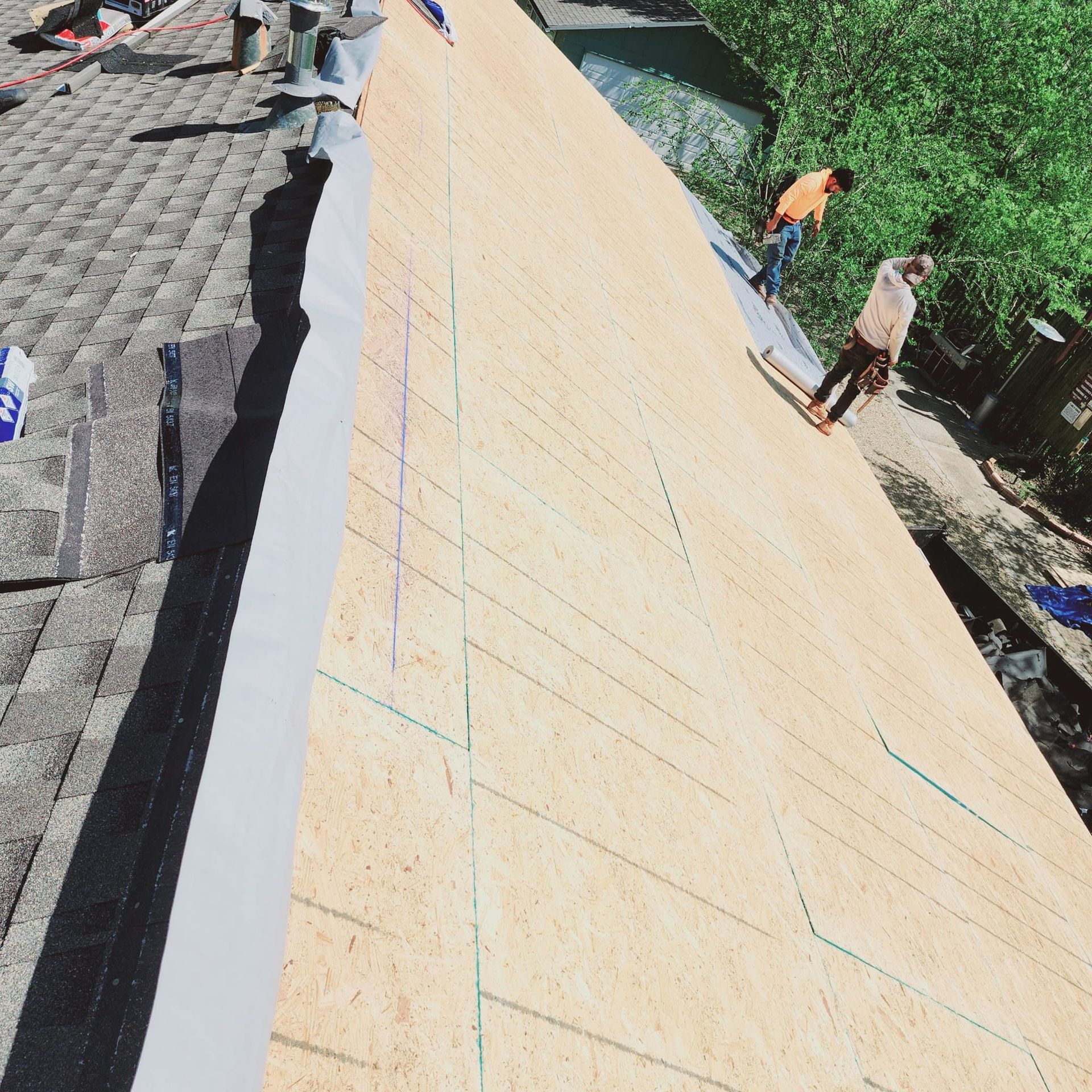 Workers installing a large tan roof under construction beside a tiled roof and green trees