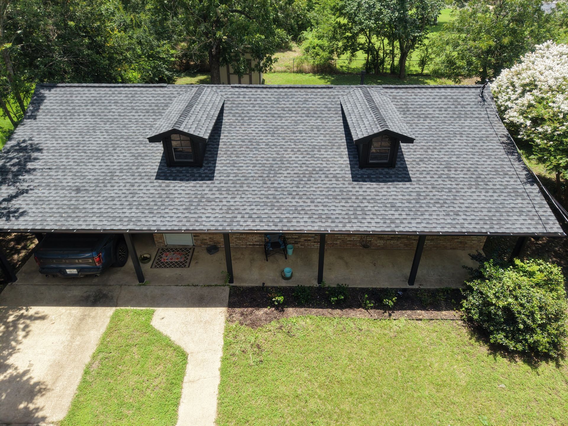 Aerial view of a gray-roofed house with two dormers, a carport, and a green front lawn.