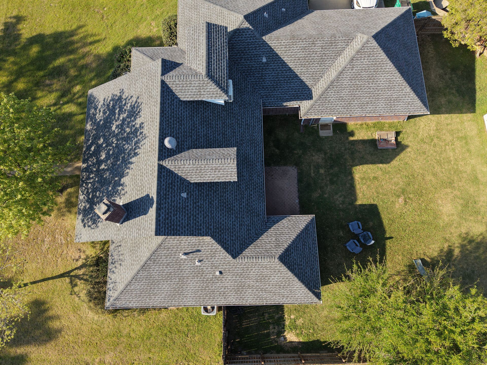 Aerial view of a gray-roofed house with a lawn and trees casting shadows.