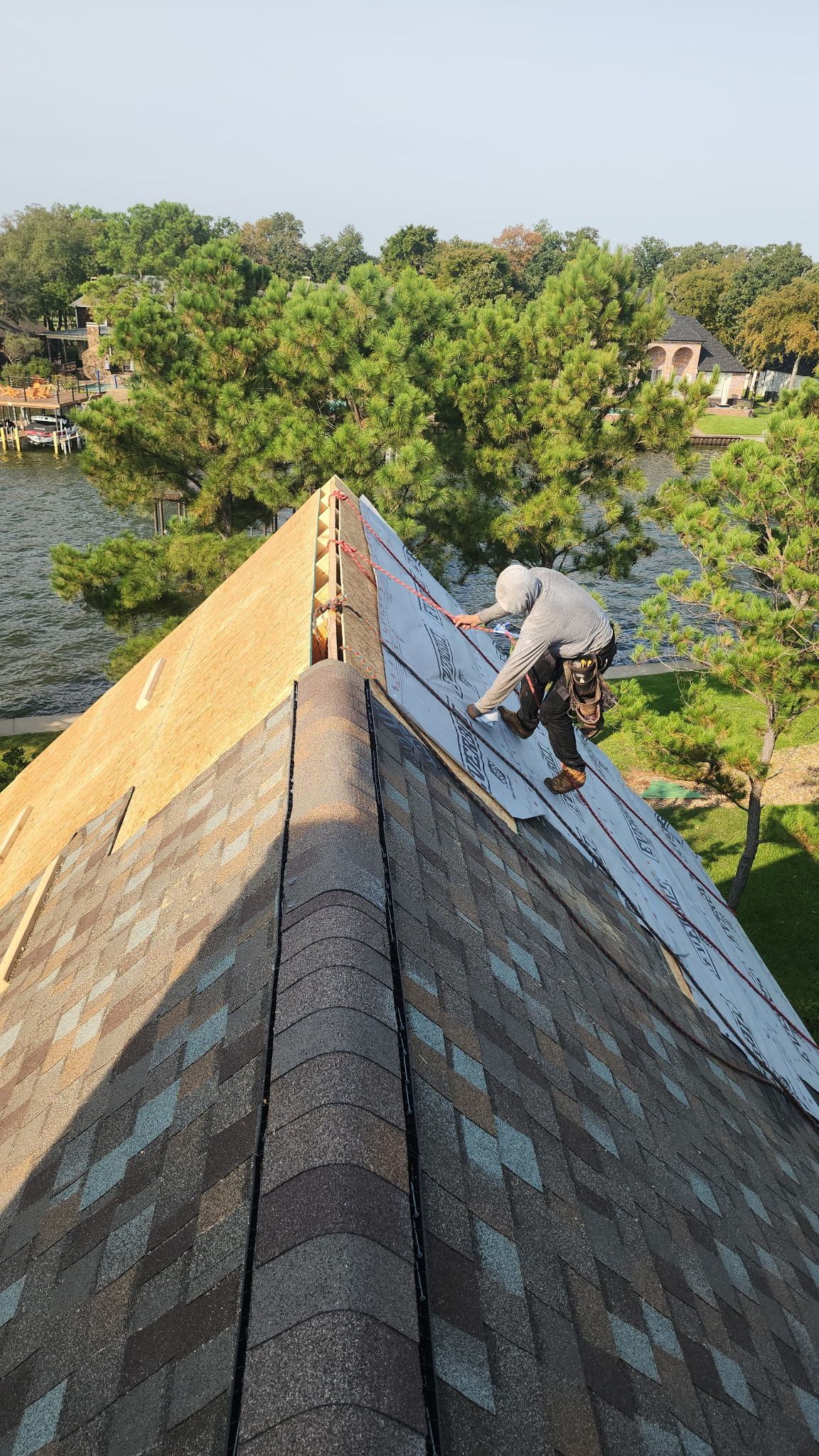 Worker on a steep shingled roof, repairing along the ridge above trees and water.