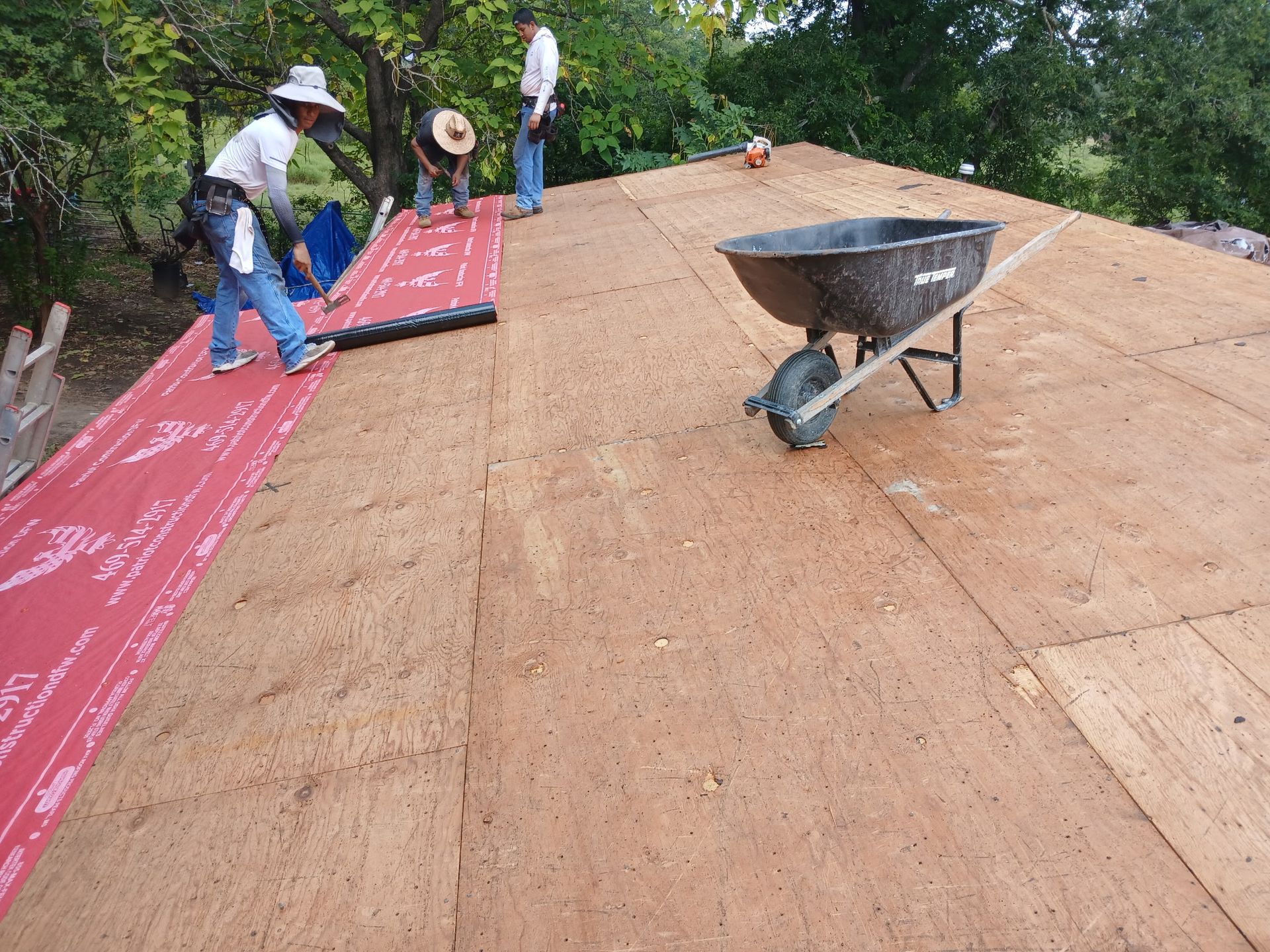 Workers installing red roofing underlayment on a sloped roof beside a wheelbarrow.