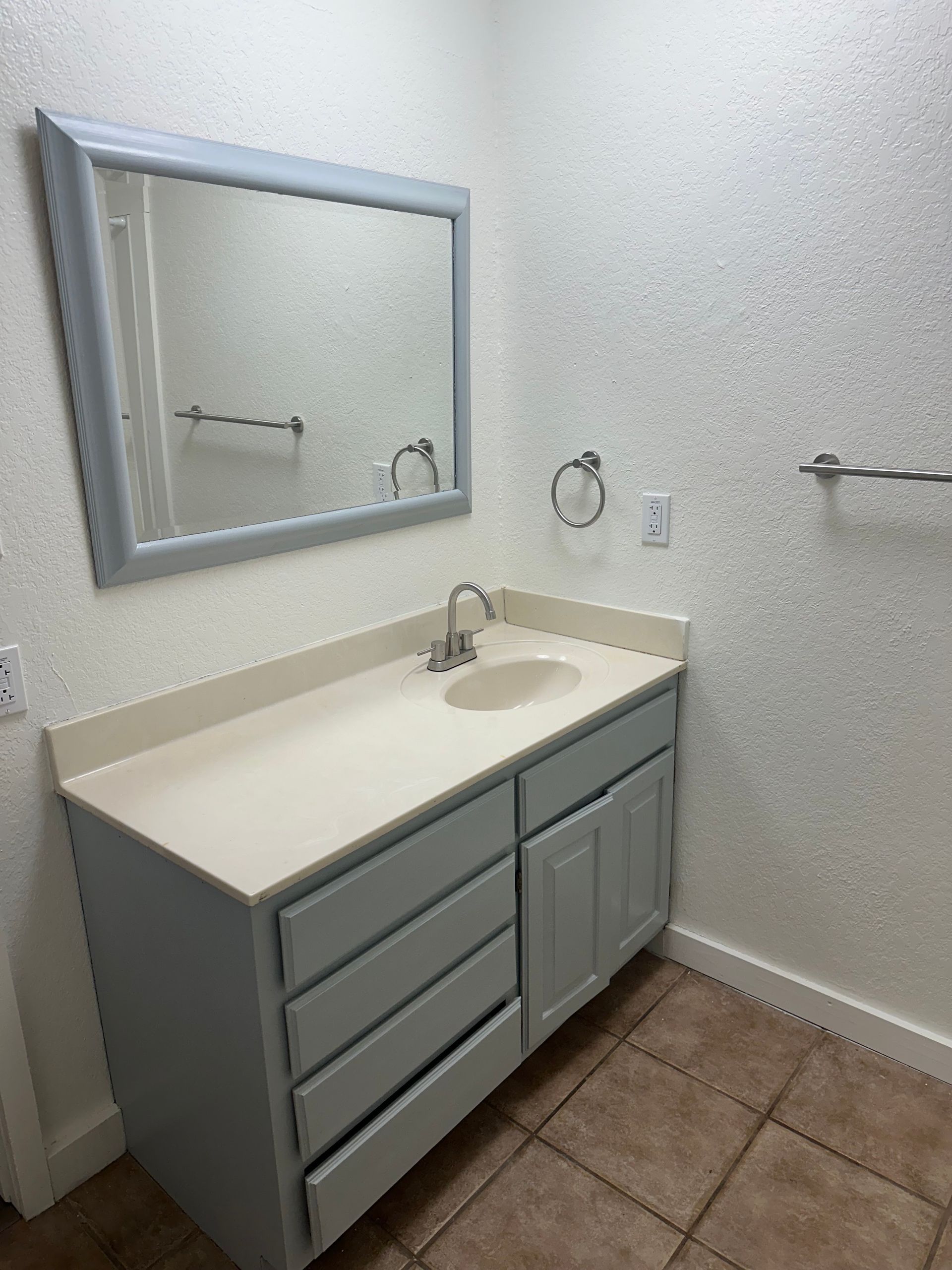 Small bathroom vanity with mirror, white sink, gray cabinets, towel ring, and tiled floor.