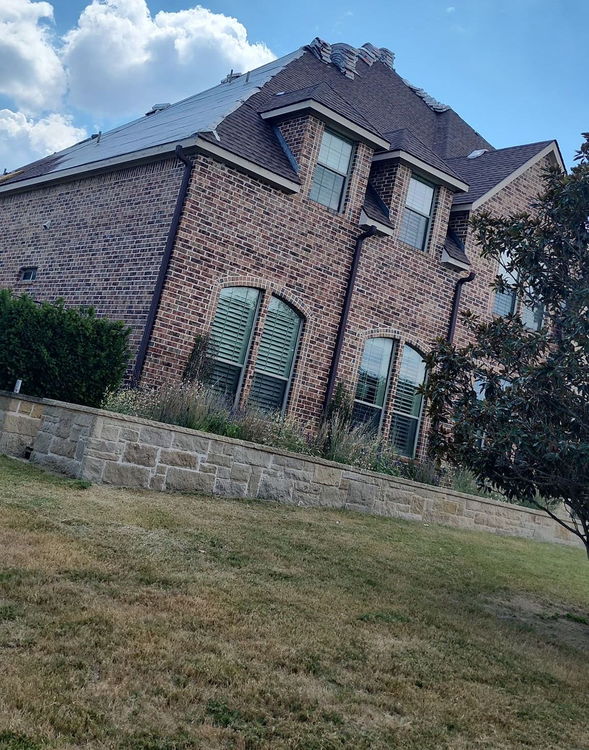 Brick house with dark roof and arched windows, viewed from a grassy yard under a cloudy blue sky