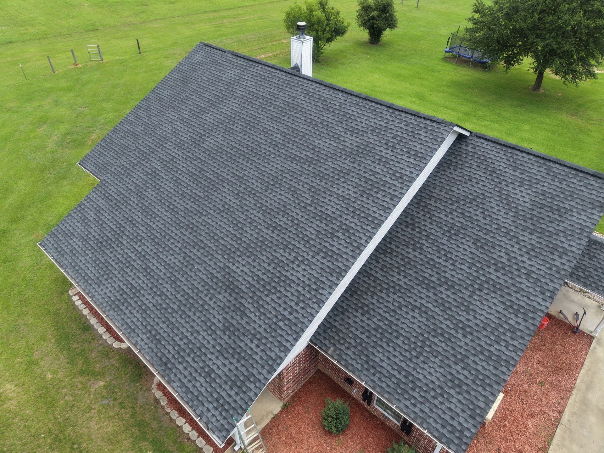 Aerial view of a house with dark gray shingle roof and red brick exterior surrounded by green lawn.