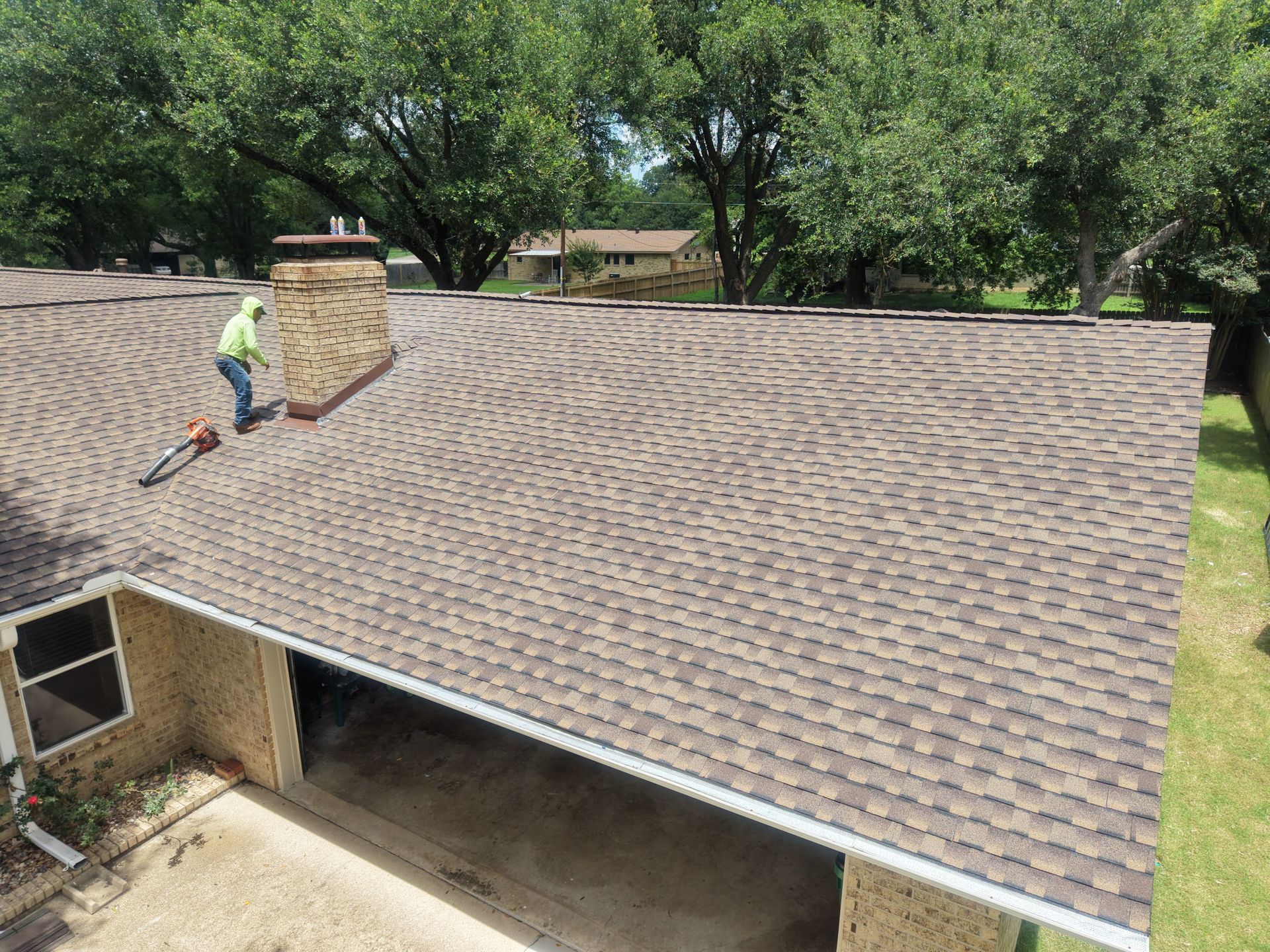 Worker on a shingled roof beside a chimney, with trees in the background.
